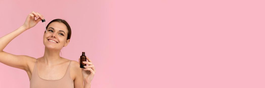 Smiling woman holding vitamin c serum near her face on pink background. Skin care and health concept. Web banner.