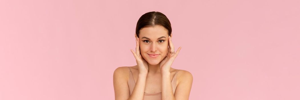 Skin care. Woman with beauty face touching healthy facial skin. Smiling female model looking at the camera and smiling over pink background. Web banner.