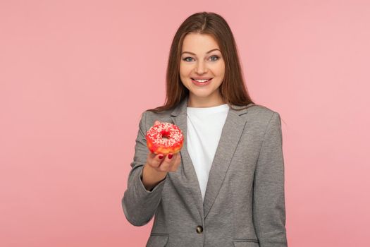 Portrait of elegant woman employee in suit jacket holding sweet doughnut and smiling to camera, showing unhealthy snack, junk food for business people. indoor studio shot isolated on pink background