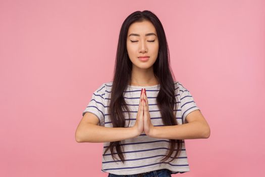 Calmness, harmony and yoga practice. Portrait of peaceful girl with long brunette hair in striped t-shirt holding arms in namaste gesture and meditating. indoor studio shot isolated on pink background