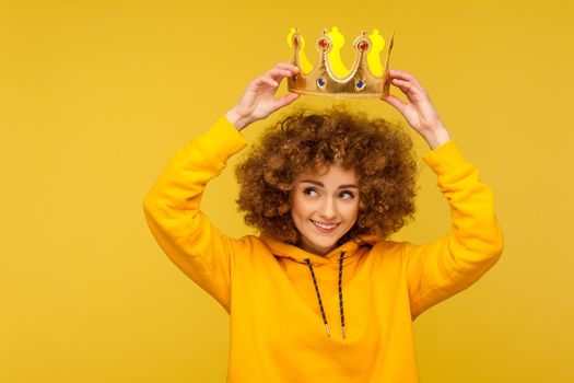I am queen. Portrait of happy curly-haired hipster woman putting crown on head and smiling, concept of self confidence in success, self-motivation and dreams to be best. indoor studio shot isolated