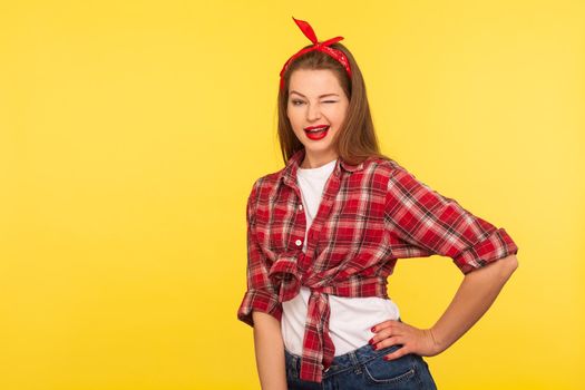 Portrait of cheerful optimistic coquette pinup girl in checkered shirt and headband winking to camera with playful flirty expression, blinking eye. retro vintage 50's style, studio shot, isolated