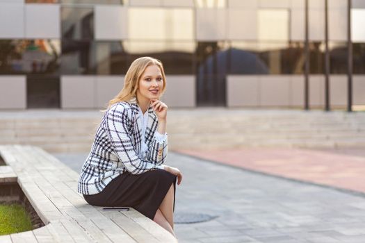 Portrait of stunning positive businesswoman with blond hair in plaid jacket and skirt sitting on bench, holding hand on chin and looking at camera with enchanting smile. outdoors, near office center