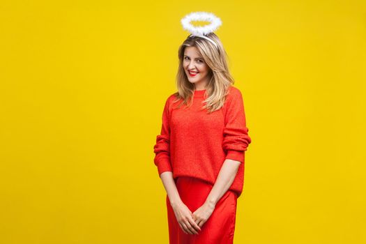 Portrait of modest angel beautiful young woman with bright makeup in red casual clothes standing with halo on her head and looking shy at camera. indoor studio shot isolated on yellow background