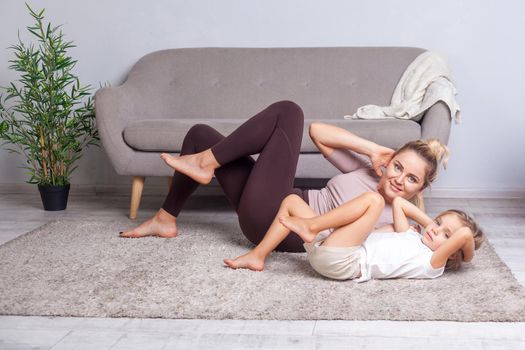 Young cheerful woman doing crunches looking at camera, mother working on abs together with her preschool daughter, happy family doing gymnastics at home, fitness training with child. body care, health