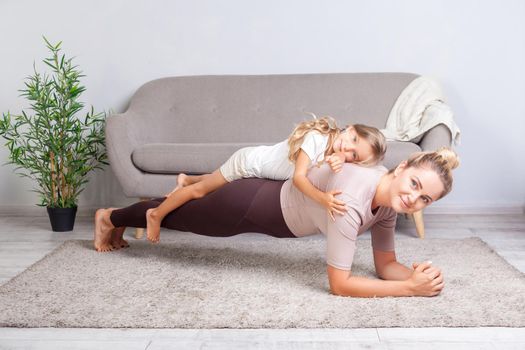 Cheerful mother doing plank workout with little daughter on her back, having fun smiling at camera, happy family doing gymnastics exercise together, fitness training with child at home. healthcare