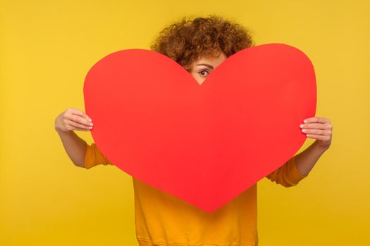 Charming young woman with fluffy curly hair holding big heart love symbol and flirting, looking playfully at camera, romance on Valentine's day. indoor studio shot isolated on yellow background