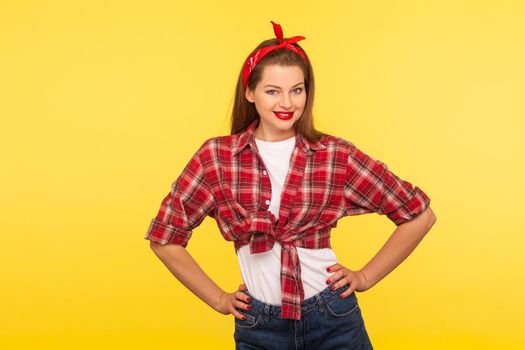 Portrait of positive pinup girl with bright makeup, red lipstick, in checkered shirt and headband, holding hands on hips and looking confident satisfied, retro vintage 50's style. studio shot isolated