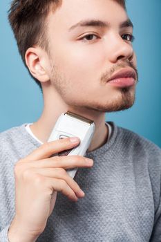 Grooming and skin care. Portrait of attentive focused brunette man with bristle and mustache holding epilator, removing facial hair, men's shaving routine. studio shot isolated on blue background