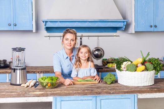 Young woman and lovely girl sitting in modern kitchen, looking at camera, mother and daughter cooking salad together, vegetarian food, fresh green vegetables and blender on table, healthy nutrition