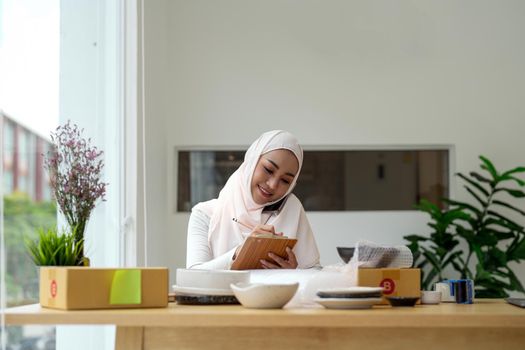 Young asian muslim woman business owner with many parcel boxes on the table happy online sales job, calling with smartphone, get an order from customers, take notes, and make arrangements for delivery by post