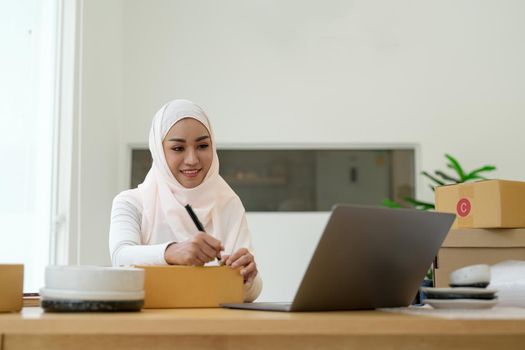 Happy young muslim woman owner writing address on cardboard box, Young Owner Woman Start up for Business Online. People with online shopping SME entrepreneur or freelance working concept