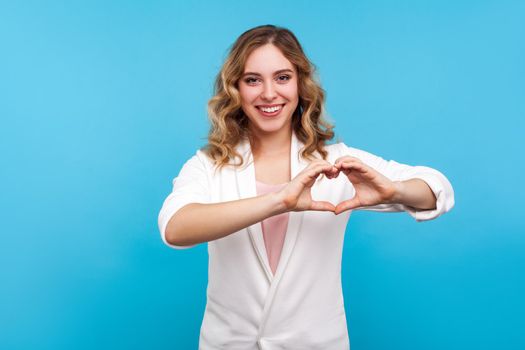 Portrait of attractive happy woman in love with wavy hair in white jacket standing, making heart shape with fingers and smiling, romance on valentine's day. studio shot isolated on blue background