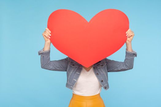 Anonymous woman in casual outfit hiding face behind huge red heart, concept of secret love fondness, unknown lover and affair, romance on Valentines day. indoor studio shot isolated on blue background