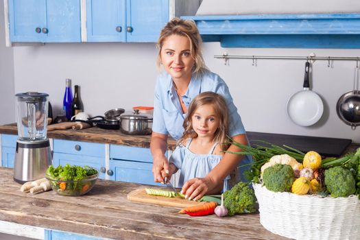 Smiling happy mother and lovely daughter cooking healthy breakfast together and looking at camera, cutting chopping vegetables, preparing vegetarian salad in kitchen, vegan food, diet nutrition
