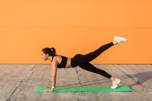 Athletic slim young woman in sportswear, black pants and top, practicing yoga, standing in Phalakasana higher plank pose with leg lift, training muscles, flexibility. Health care, sport activity