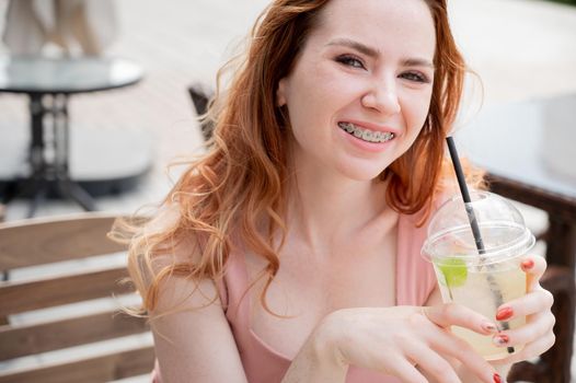 Young beautiful red-haired woman with braces drinks cooling lemonade outdoors in summer. Portrait of a smiling girl with freckles