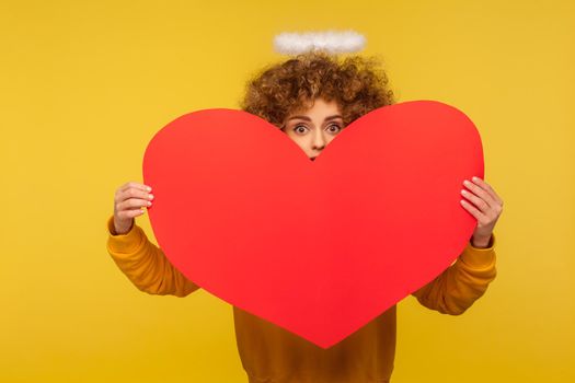 Portrait of surprised angelic curly-haired woman with saint nimbus peeking out of big red heart, her big eyes expressing amazement, shocked expression. indoor studio shot isolated on yellow background