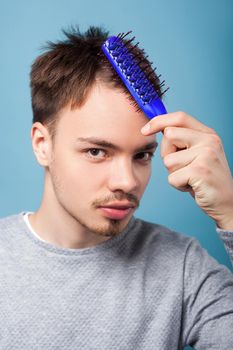 Healthy hair. Portrait of brunette man with small beard and mustache in casual sweater combing his hair, showing clean scalp skin without dandruff. indoor studio shot isolated on blue background