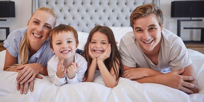 Portrait of a cheerful family with two children lying together on a bed. Little boy and girl lying on their parents bed and smiling at the camera. Caucasian couple bonding with their son and daughter. Siblings enjoying free time with their mother and father.