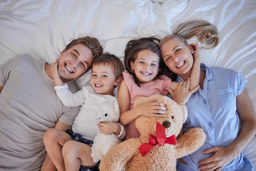 Portrait of a happy caucasian family with two children relaxing and lying together on a bed at home, from above. Little brother and sister holding stuffed animals and touching mom and dads face.