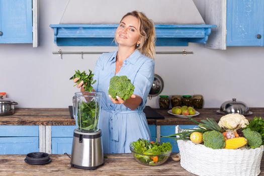 Cheerful woman holding green vegetables and putting ingredients into blender, smiling at camera, fit girl preparing healthy smoothie for breakfast, cooking salad in modern kitchen, vegetarian diet