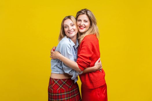 Best friends hugging. Portrait of two charming women in stylish casual clothes standing, embracing together and looking at camera with toothy smile. indoor studio shot isolated on yellow background