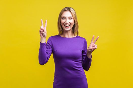 Portrait of joyous beautiful woman in elegant purple dress smiling at camera and showing victory gesture with both hands, winner excited about success. indoor studio shot isolated on yellow background