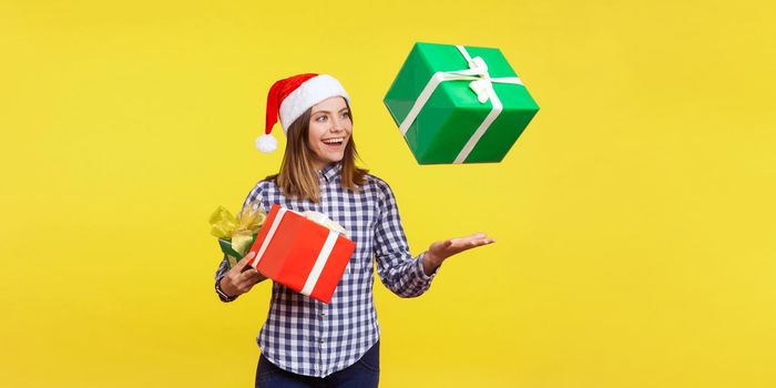 Portrait of happy brunette young woman in santa hat and checkered shirt standing throwing gift boxes in air or catching presents, christmas shopping. indoor studio shot isolated on yellow background