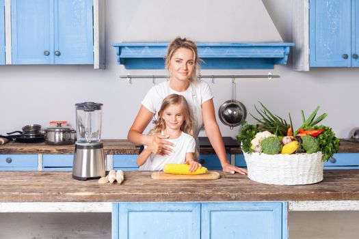 Happy attractive mother hugging daughter, standing together in kitchen with modern furniture, little girl holding corn, basket of fresh vegetables on table, vegan nutrition, healthy food. indoor