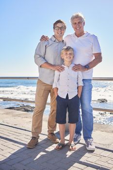 Male family members posing together at the beach on a sunny day. Grandfather, father and grandson standing together on seaside promenade. Multi-generation family of men and little boy spending time together.