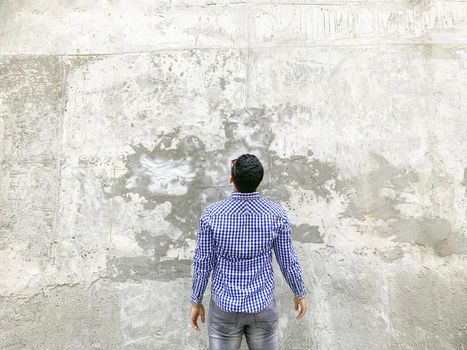 Backside of young man in checkered blue shirt and sunglasses standing against concrete gray wall. looking at background empty copyspace.