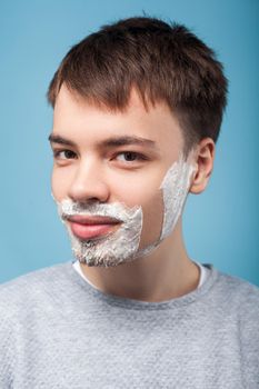 Portrait of happy young man in casual clothes looking aside with shaving foam on his bristle, satisfied with soft skin on shaved part of his cheek. indoor studio shot isolated on blue background