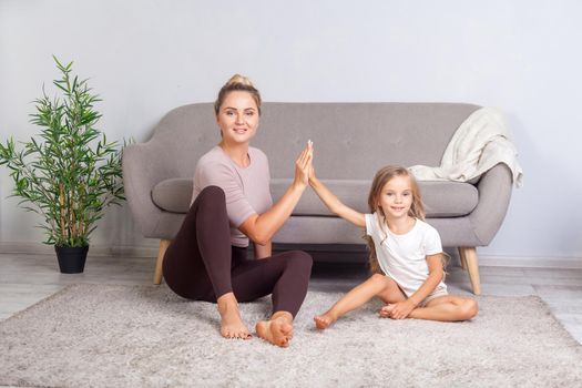 Fit attractive woman giving high five to happy little daughter, teamwork concept, mother and child sitting on floor taking break relaxing after sport fitness, resting together. trust to teammate