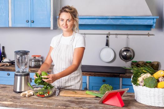 Attractive young woman in apron cooking green salad, preparing vegetarian food in modern kitchen and looking at camera, blender and basket of fresh vegetables on table, vegan diet, healthy nutrition