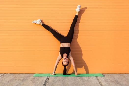 Overjoyed excited girl with perfect athletic body in tight sportswear doing yoga handstand pose against wall and laughing, shouting from happiness. Gymnastics for body balance outdoor workouts