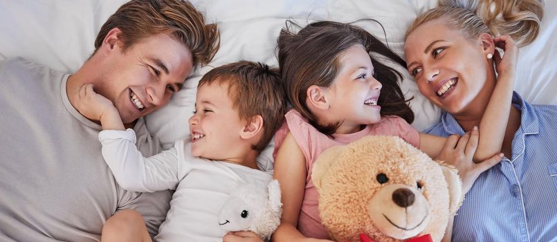 Close up of a happy caucasian family with two children relaxing and lying together on a bed at home, from above. Little brother and sister holding stuffed animals and touching mom and dads face while bonding at home.