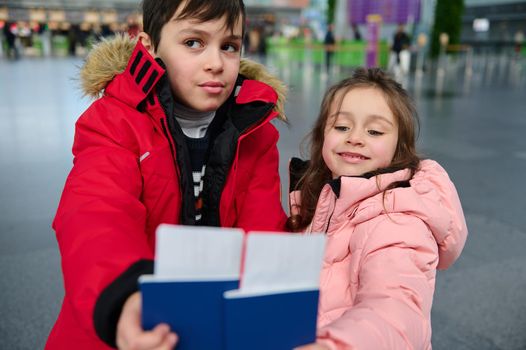 Adorable Caucasian kids- boy and girl, siblings holding boarding pass and Identity document in outstretched hands while waiting for check-in flight in the departure hall of an international airport