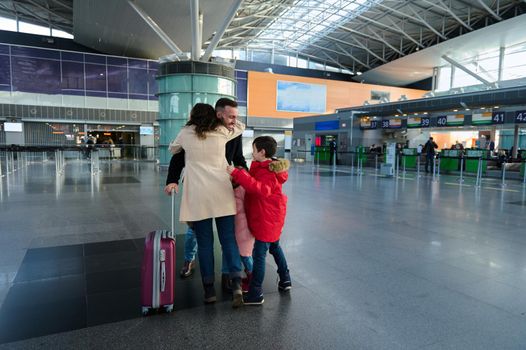 Happy young family embracing at the airport. Husband hugs his wife and children after returning from a business trip in the arrival terminal of the international airport. Long-awaited family reuniting