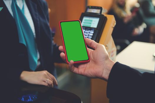 Man's hand holding smartphone with green chroma key blank screen with copy space for mobile apps and advertising, near bank terminal while going through flight check-in board at airport. Close-up