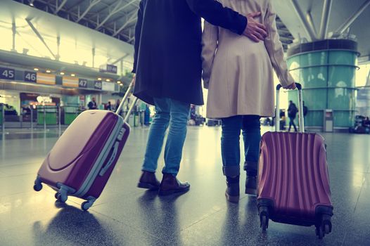 Close-up of young couple with suitcases walking along the departure hall of international airport on the blurred background of flight check-in counters. Back view man hugging woman in arrival terminal
