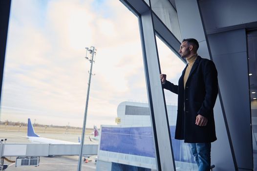 Full length portrait of a confident man passenger looking through panoramic windows overlooking the runway in the departure terminal of an international airport while waiting for a flight boarding