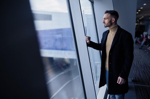 Pensive handsome young man, business traveler, transit passenger standing by panoramic windows overlooking runway in the departure terminal of an International Airport, awaiting the flight boarding