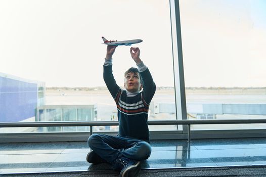 Adorable 10 years old boy plays with a toy plane sitting on the floor against the background of panoramic windows overlooking runway with aircraft in the departure terminal of international airport