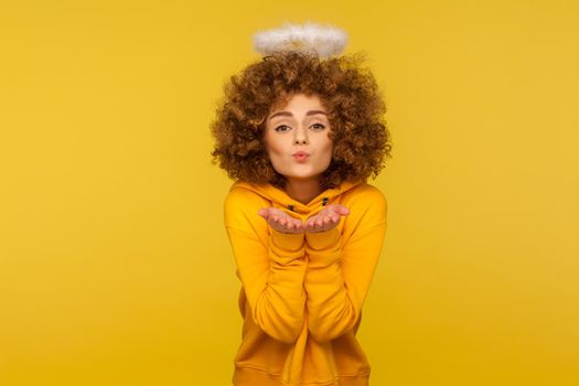 Portrait of kind-hearted curly-haired hipster woman with saint nimbus sending air kiss over palms, demonstrating romantic feelings, angelic love. indoor studio shot isolated on yellow background