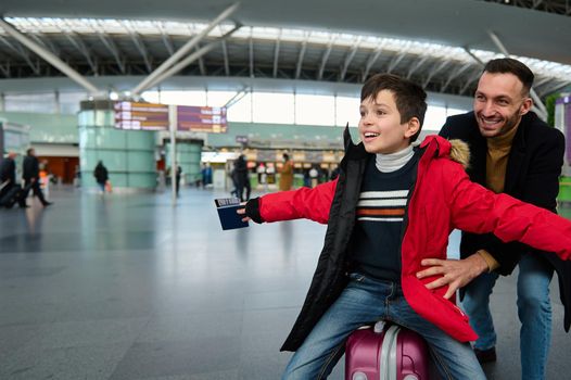 Cheerful happy young dad rides his son on a suitcase in the departure hall of the international airport. Dad and son having fun together while waiting for passport and customs control and boarding