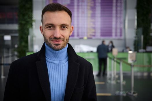 Handsome Caucasian young man in casual wear, traveler, passenger stands at flight information board with timetable in international airport arrival or departure terminal, confidently looking at camera
