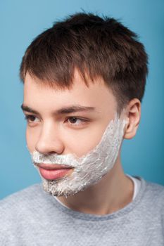 Portrait of young man in casual sweater looking aside with shaving foam on his bristle and mustache, ready to shave beard, hygiene and skincare concept. indoor studio shot isolated on blue background