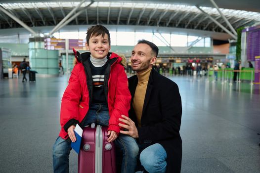 Handsome adorable preschooler, Caucasian child boy in warm red down jacket holding boarding pass and passport sitting on suitcase next to his loving father in international airport departure terminal