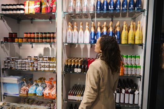 Rear view of a charming brunette traveler woman transit passenger looking at stalls with different food items and various goods while shopping in a duty free shop in the international airport